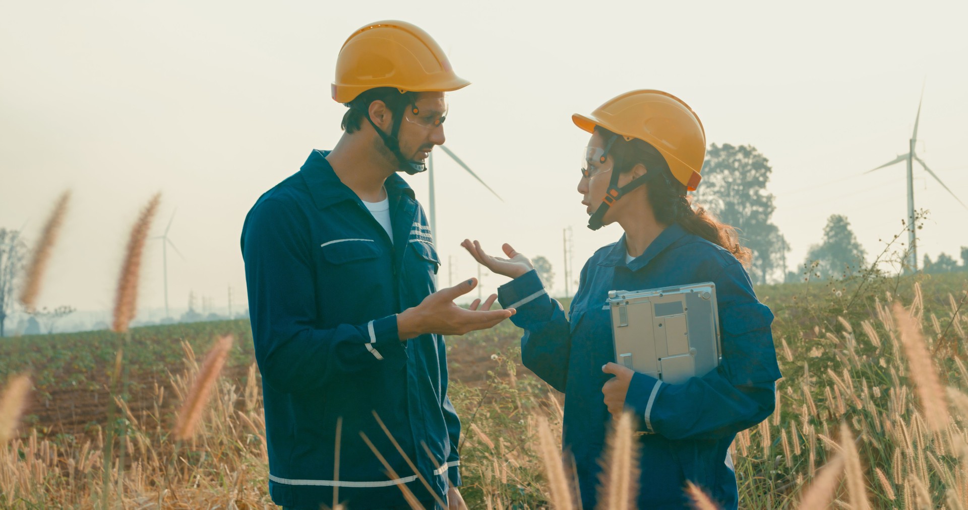Technicians review technical data on laptop while conducting maintenance checks at wind turbine site in vast agricultural field. Green ecological power generation wind sustainable energy.
